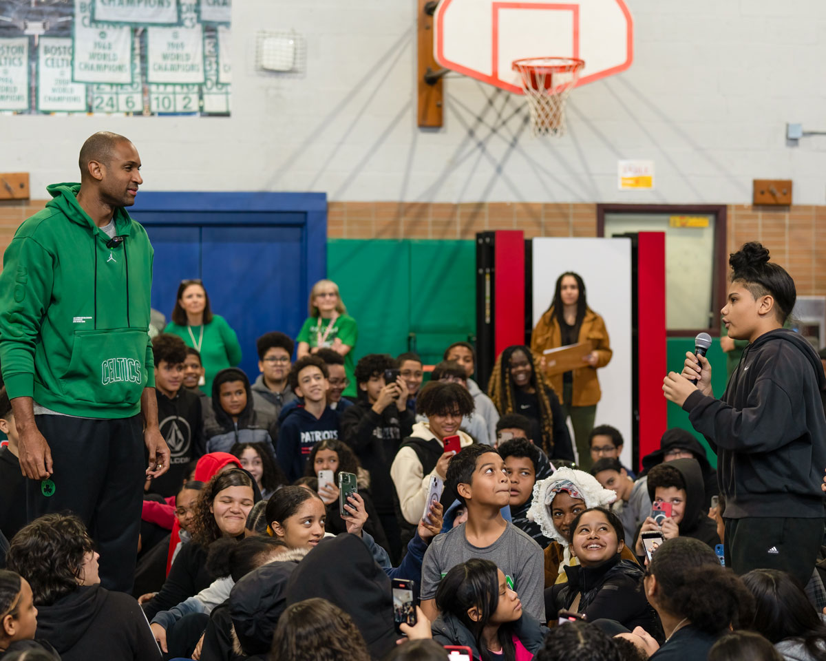 Celtic player and a student standing in the middle of a crowd of students that is sitting down